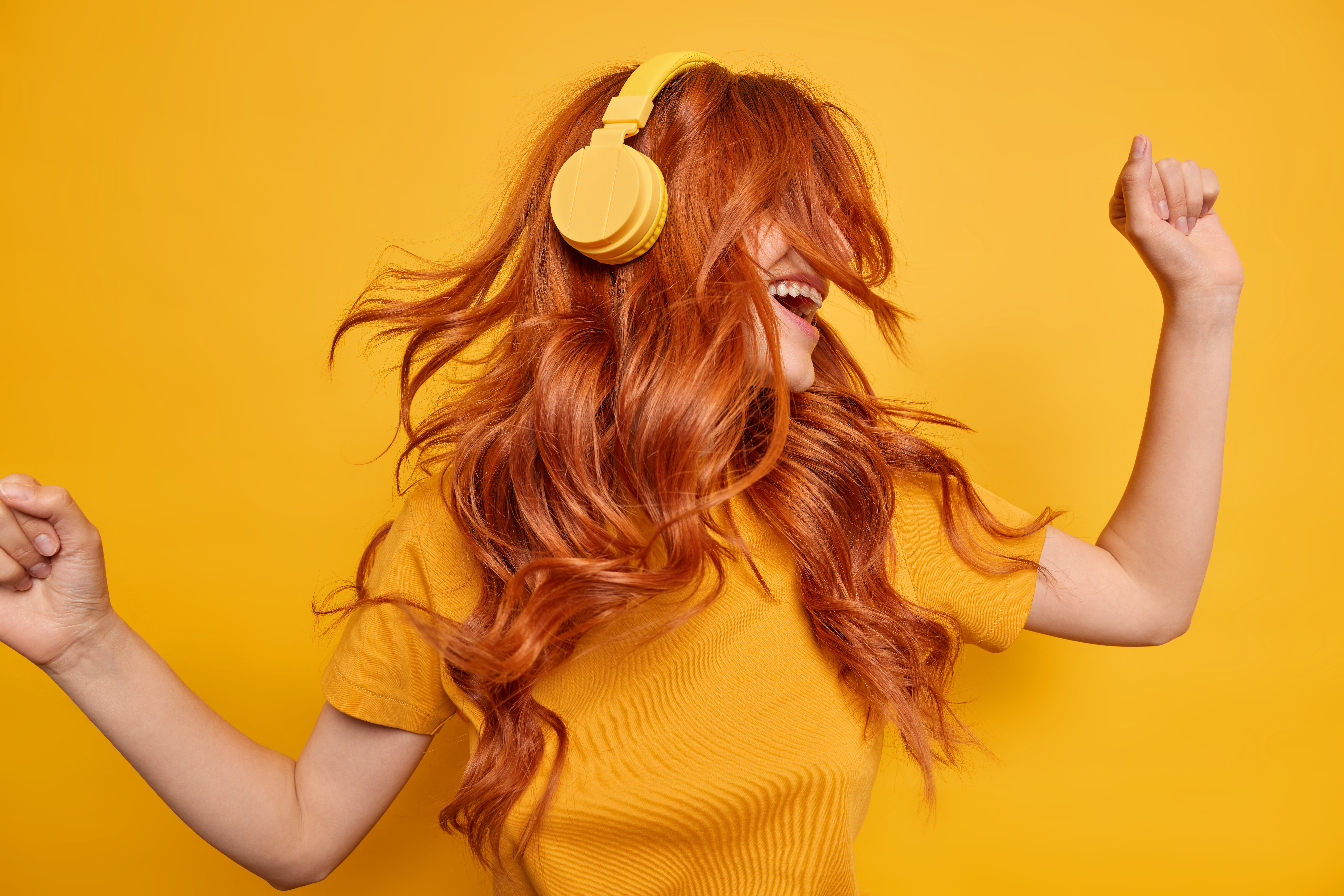 cool millennial girl keeps arms raised dances carefree enjoys every bit of music wears wireless headphones on ears has red hair floating on wind dressed in casual t shirt isolated over yellow wall